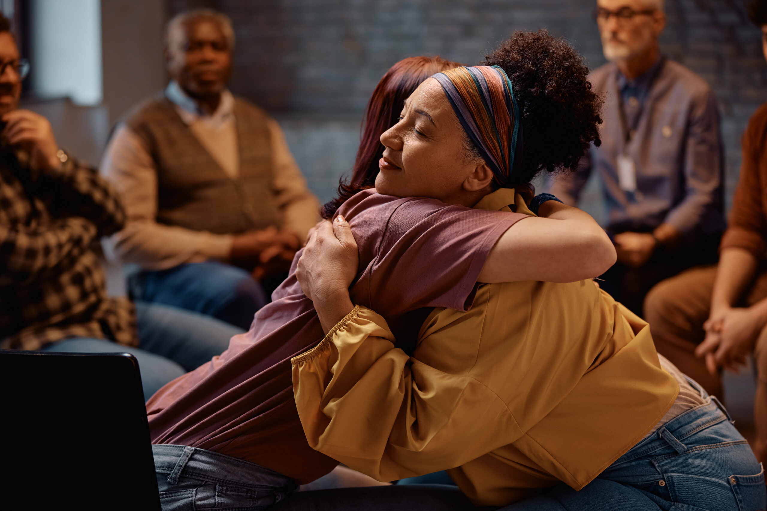 Two women embrace in a supportive group setting, symbolizing emotional resilience, healing, and the power of connection in building a happier, healthier life.